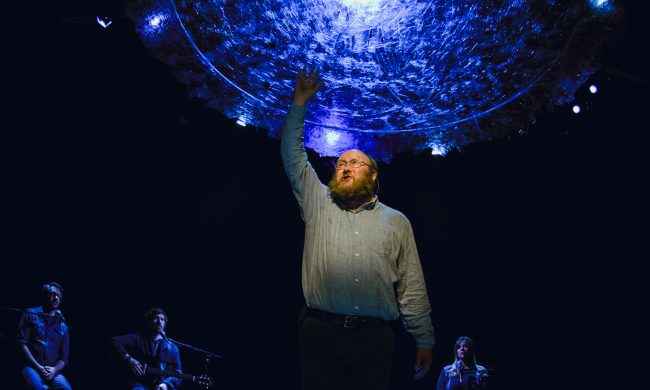 Photo from a past Artistic Fraud presentation of a man on stage standing on a chair reaching towards the ceiling.