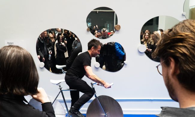 Photo of a man in a black t-shirt and black pants riding a stationary bike in front of a group of onlookers.