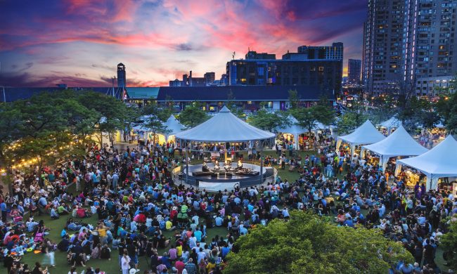 Image of patrons gathered around a round stage with white canopy surrounded by white vendor tents and lush greenery, set against a pink and purple summer sunset