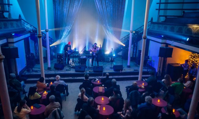 Photo of an indoor event with 2 musicians performing on a stage and audience seated at tables.