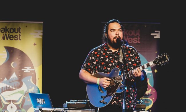 Musician with a blue guitar and a microphone in front of them. The have dark black hair that is pulled back and a beard. They are wearing a button up shirt with polka dots and pink and red hearts. There is table behind them with a laptop and a turntable. Two BreakOut West pop up banners are behind the table.
