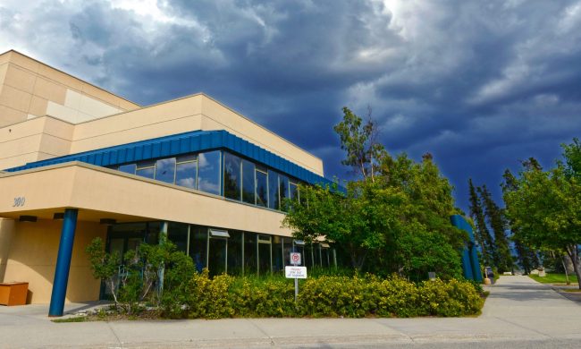 Exterior of the Yukon Arts Centre, a building made of beige concrete, blue pillars and detailing with lots of windows surrounded by green trees and bushes. The sky in the background is cloudy and dark blue.