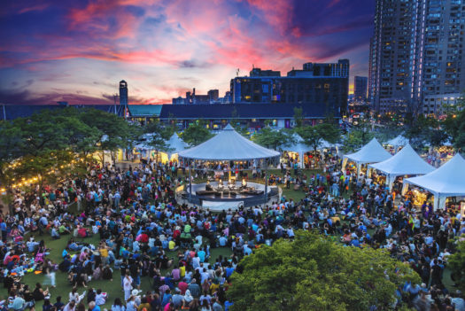 People gathered around a quartet of artists performing on a round stage with a white conical canopy surrounded by white vendor tents and greenery with a pink and orange sunset in the background.