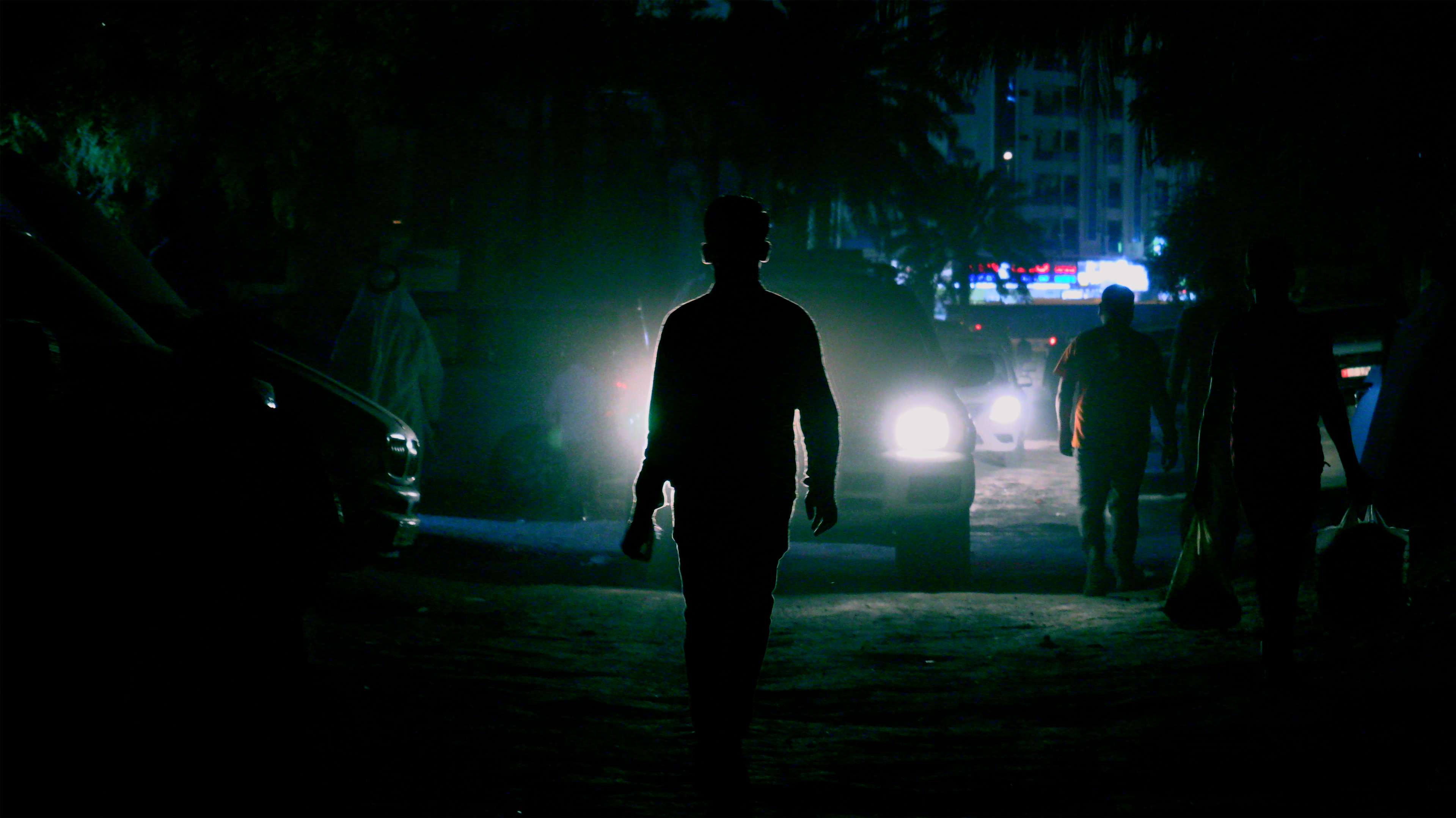 Silhouette of a person walking down a road with a car coming towards them at night. In the background are palm trees, a building and other pedestrians.