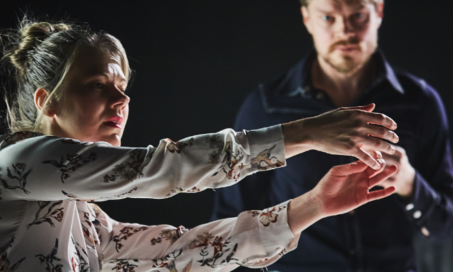 A dancer standing sideways on-stage with hair pulled into a bun and their arms reaching out. In the blurred background is another dancer walking towards them.