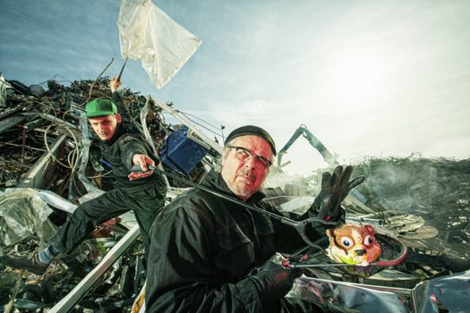 Two performers wearing black long-sleeve jumpsuits moving across a pile of garbage at an outdoor landfill in daytime.
