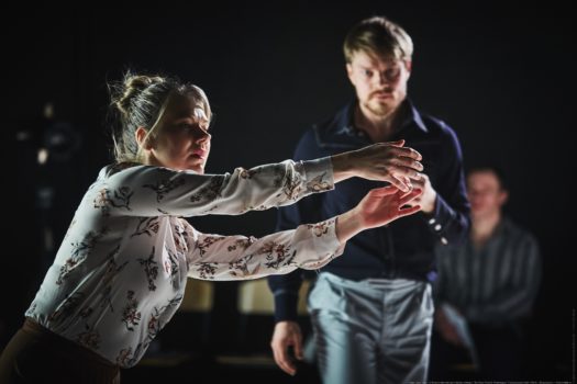 Dancer Maria Nurmela standing sideways on-stage with hair pulled into a bun and arms reaching out. In the blurred background dancer Ville Oinonen walks towards her.