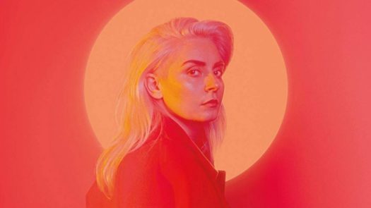 Music artist Eivør, a woman with long blonde hair wearing a black top poses in front of a yellow circle surrounded by soft red light.
