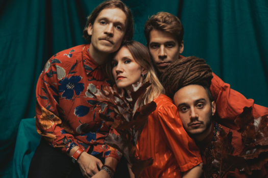 Music quartet Kolonien pose leaning on each other with soft facial expressions, wearing flowing red shirts and holding a branch of leaves in front of a teal green satin backdrop.