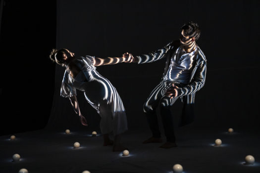 Two performers in loose white costumes lean away from each other and counterbalance by holding the opposite hand of the other. The stage is dark with white striped lighting on the performers and small white spheres placed around them on the floor.