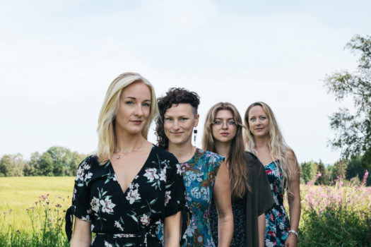 Four members of the band Kongero stand in a field in a diagonal line facing the camera each wearing a floral dress in shades of black, white and blue during daytime.