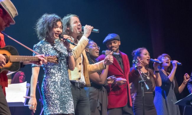 Six singers standing in a line on an indoor stage while performing with a guitar player on each side of them against a black backdrop.
