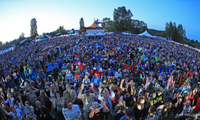 Aerial view of a crowded audience at an outdoor music festival at dusk with white vendor tents and trees in the background.