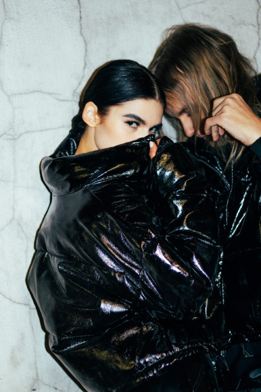 Music duo Kitka face each other while pulling up the collars of their shiny black puffer jackets with one of them peaking at the camera.