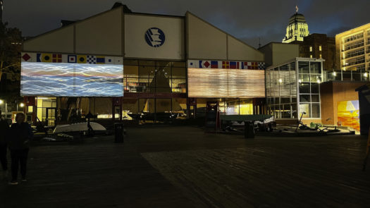 A colourful video installation of sea water projected onto the outside of a building with people walking by at dusk.