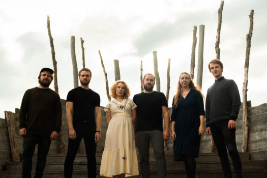 Six members of VÍÍK stand side by side outside in front of a wooden construction all wearing black clothing except for one person left of centre wearing a white v-neck short sleeve dress.