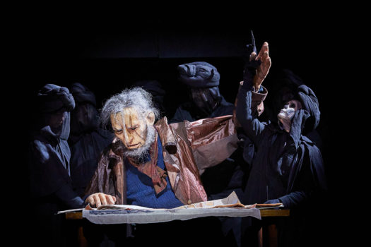 A performer playing Moby Dick sits on-stage at a table with props on it with their right arm being raised by one of five people behind them wearing black robes and headwraps with skeletal facial makeup.