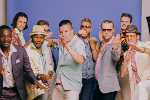 Group shot of nine members of Helsinki Cotonou Ensemble with expressive facial expressions and poses against a blue backdrop.